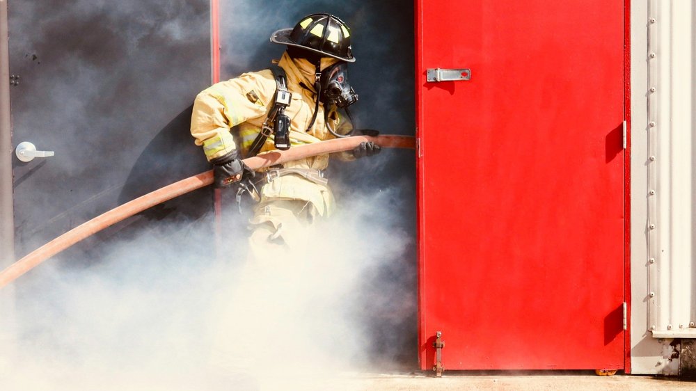 A firefighter in full gear pulls a hose through smoke beside an open red door.