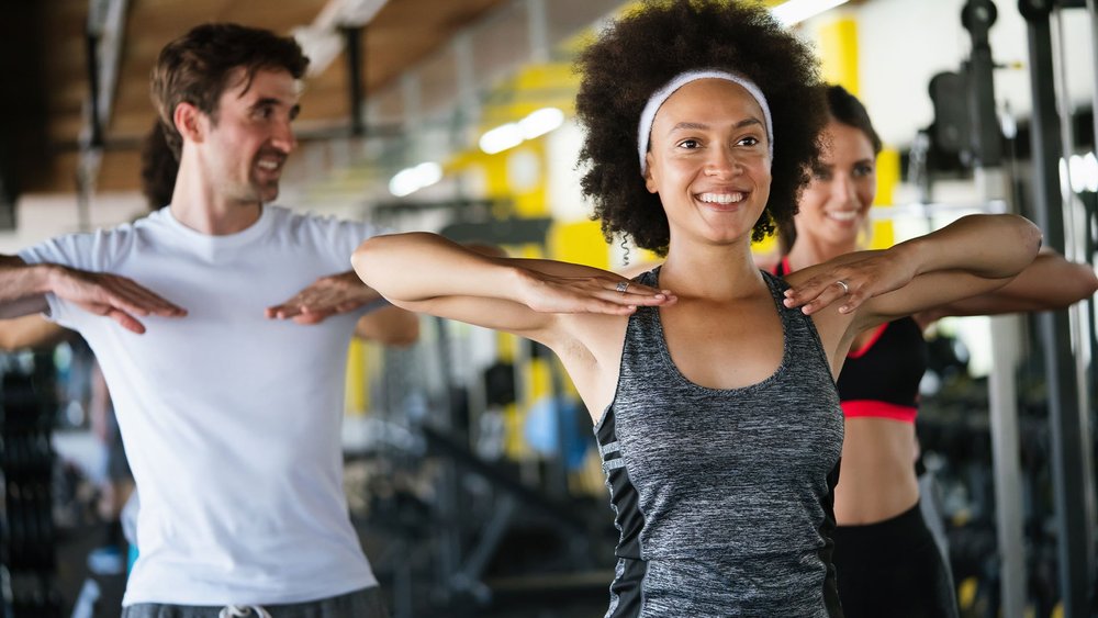 Three people do a shoulder stretch together in a gym, smiling.