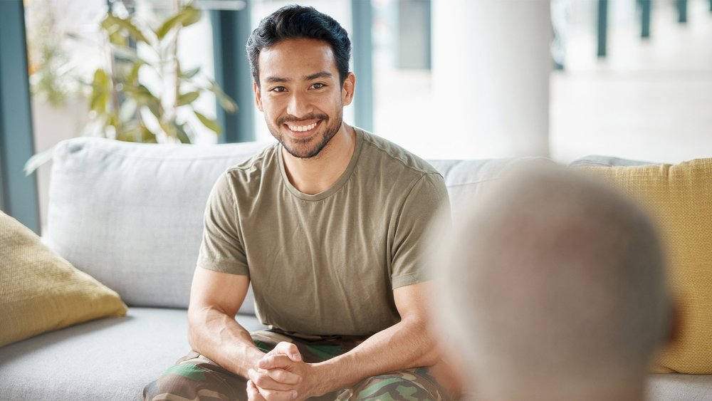 A smiling man sits on a couch talking with another person nearby.