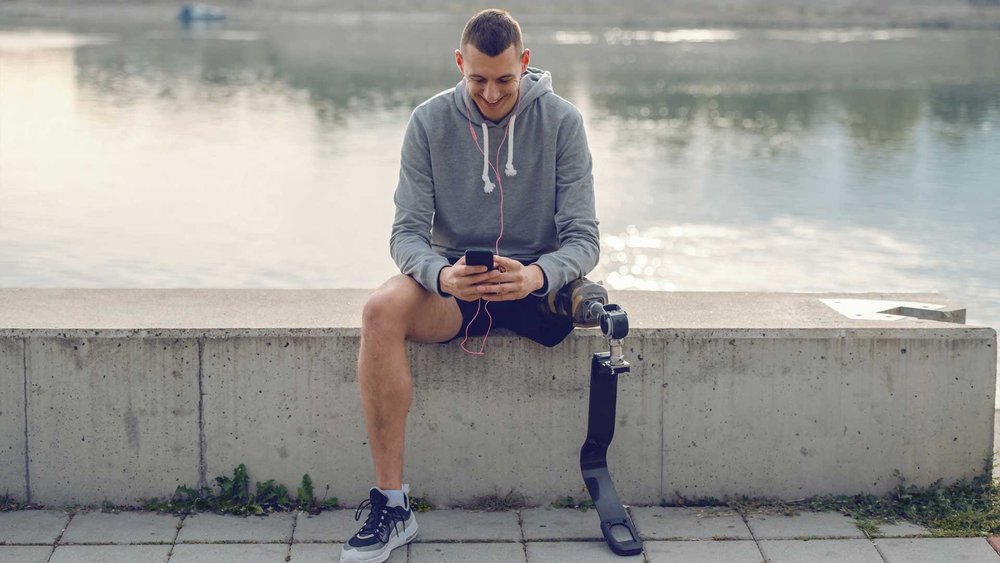A man with a leg prosthesis sits on a wall by the water, looking at his phone.