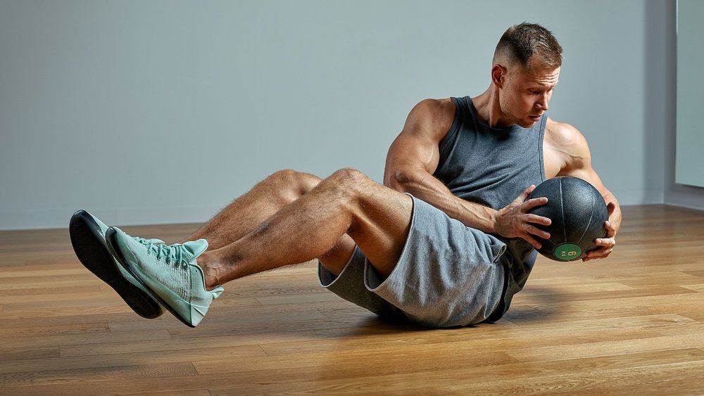 A shirtless man performs a seated Russian twist exercise holding a medicine ball.