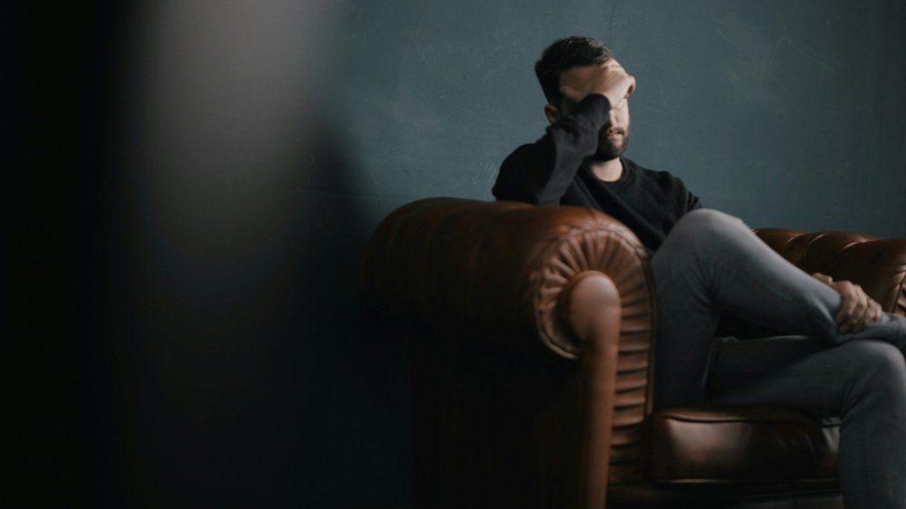 A man sits on a leather couch with his head in his hand, looking troubled.