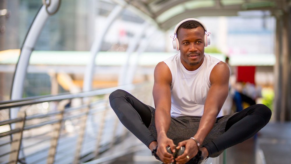 A man wearing headphones sits cross-legged on a walkway, stretching toward his feet.