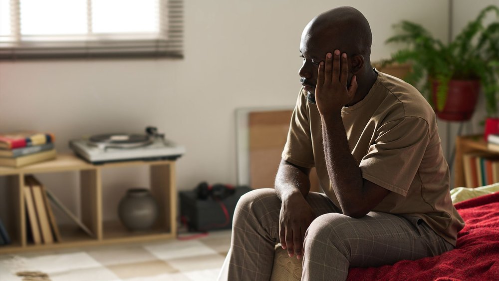 A man sits on the edge of a bed with a hand over his face.