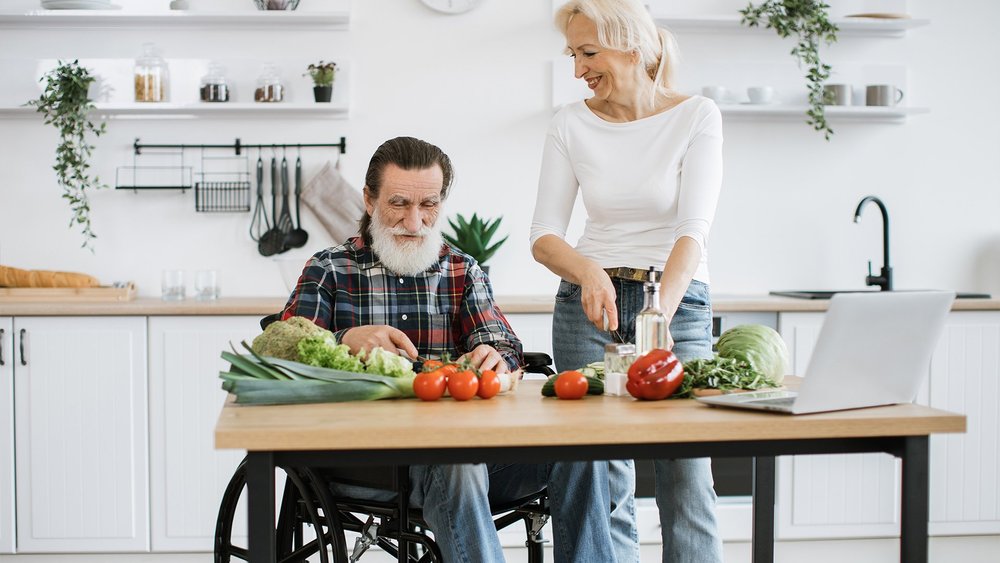 A man in a wheelchair at a kitchen table spread with fresh vegetables while a woman prepares food.