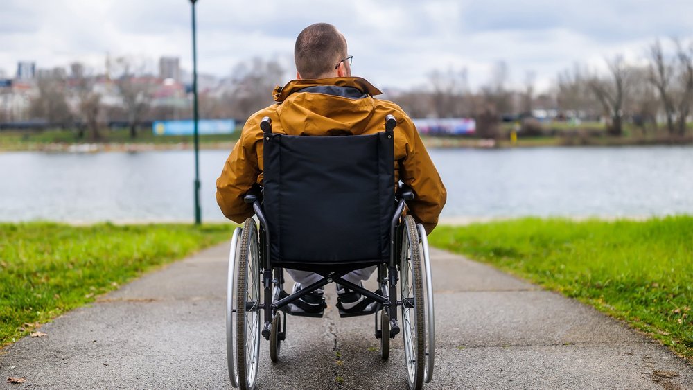 A man in a wheelchair, seen from behind, faces a lake with his arms resting on the wheels.