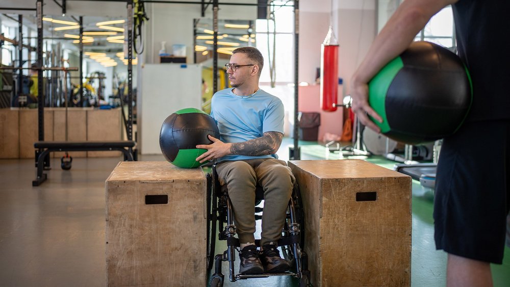 A man in a wheelchair holds a medicine ball between two plyo boxes as a partner watches.
