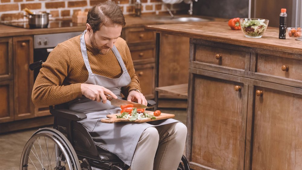 A man in a wheelchair wears an apron and chops vegetables in a kitchen.