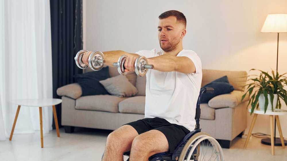 A man in a wheelchair lifts dumbbells with both arms in a living room.
