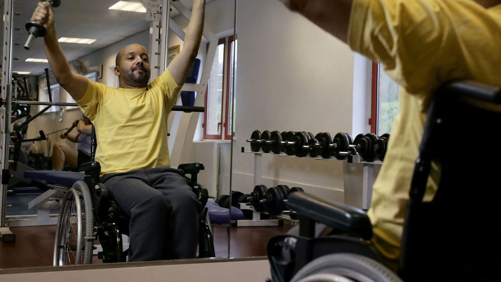 A man in a wheelchair lifts a dumbbell overhead in front of a gym mirror.