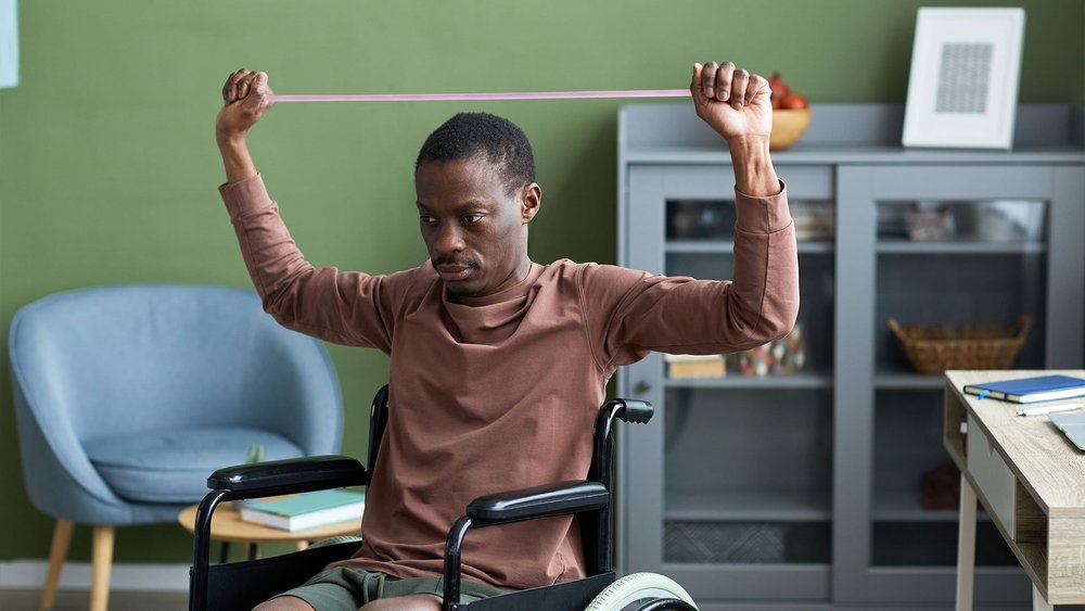 A man in a wheelchair stretches a resistance band overhead with both arms.