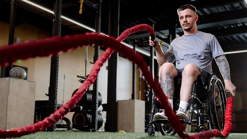 A man in a wheelchair trains with battle ropes in a gym.