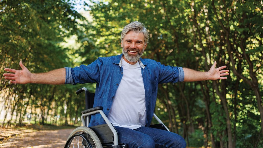 A smiling man in a wheelchair spreads his arms wide on a tree-lined path.