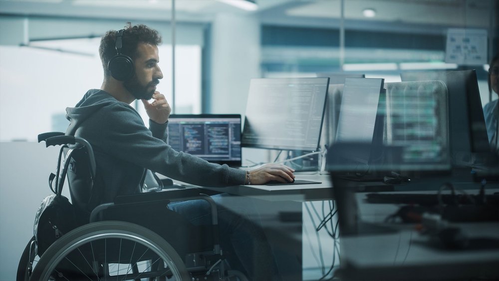 A man in a wheelchair wearing headphones works at a desk with multiple monitors.