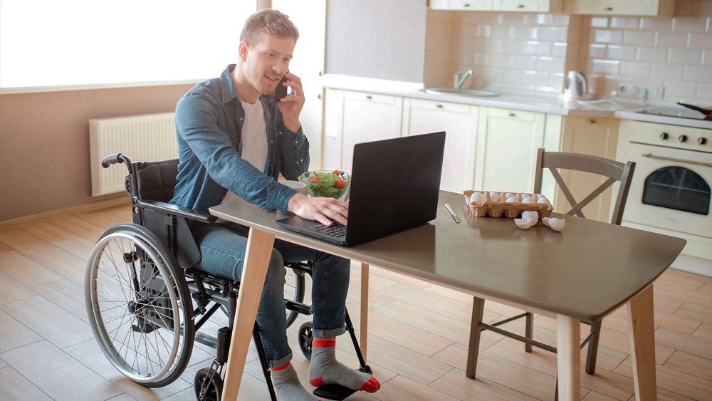 A man in a wheelchair talks on the phone while typing on a laptop at a table.