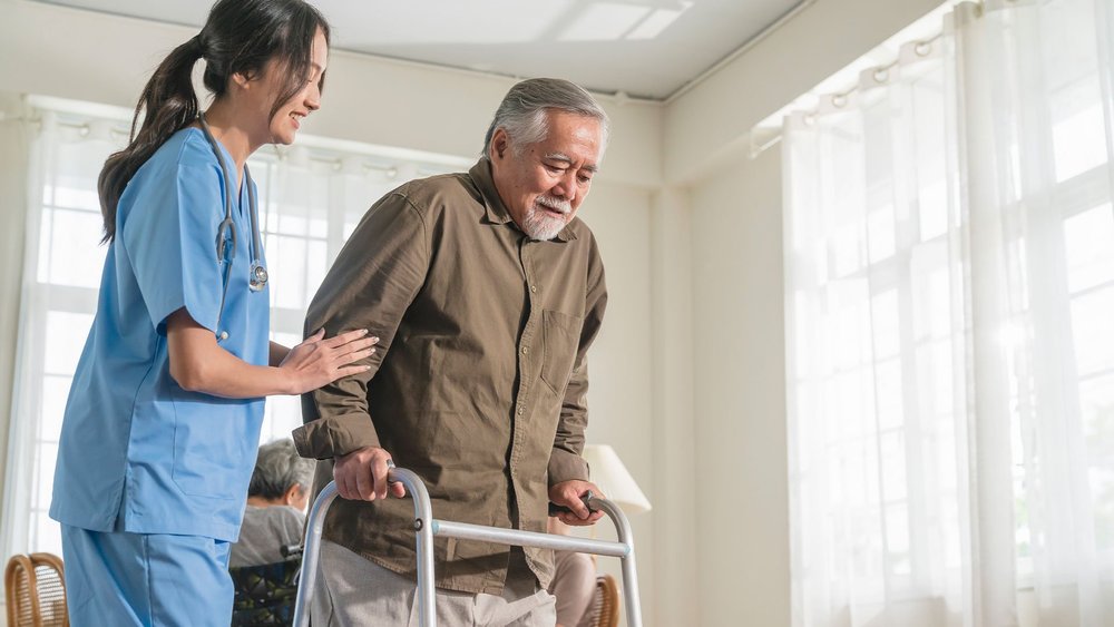 A nurse helps an older man walk using a walker in a bright room.