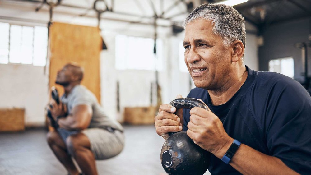 A man holds a kettlebell at the gym while another man squats in the background.