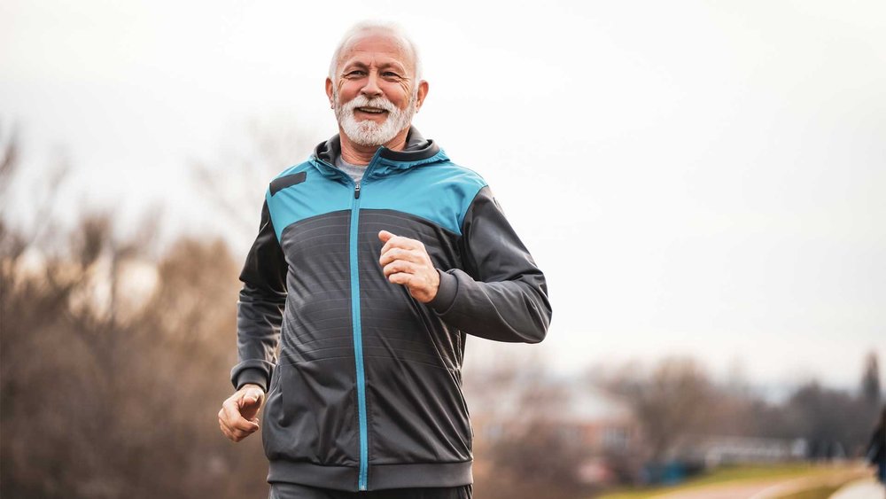An older man in a tracksuit jogs outdoors, smiling.