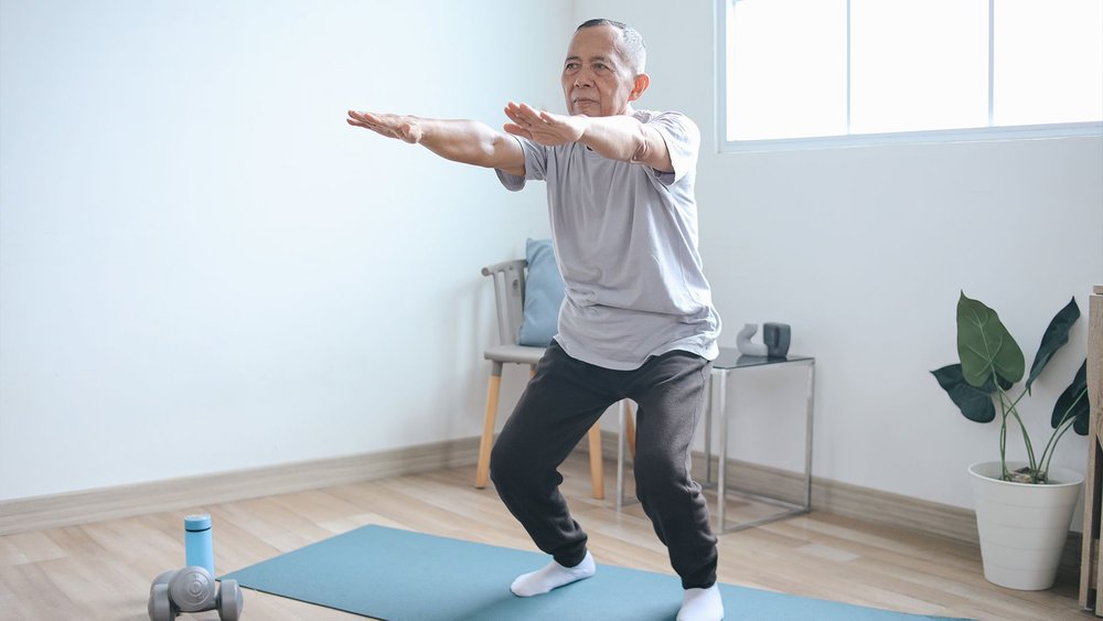 An older man balances on one leg with arms extended during a home workout.