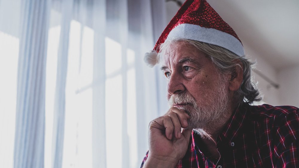 An older man in a Santa hat looks out of a bright window.