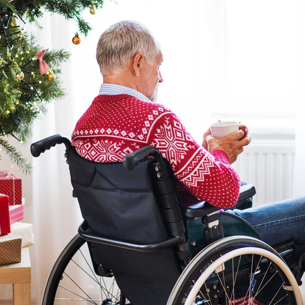 An elderly man in a wheelchair holds a mug near a decorated Christmas tree.
