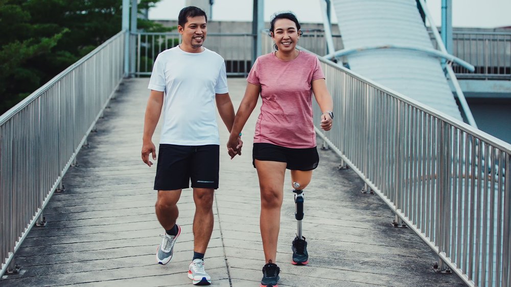 A couple holds hands while walking on a bridge, the woman wearing a prosthetic leg.