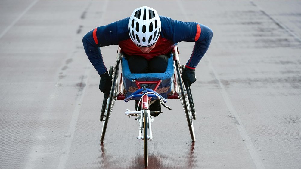 A racing wheelchair athlete in a helmet leans forward on a track.