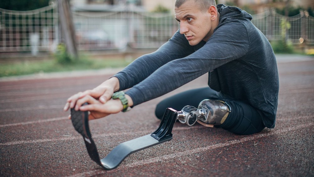 A man with a running blade prosthesis sits on a track, stretching toward his foot.