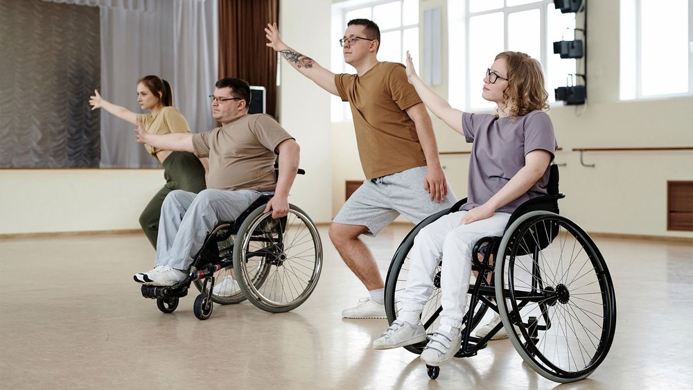 Four wheelchair users stretch their arms together in unison in a dance studio.