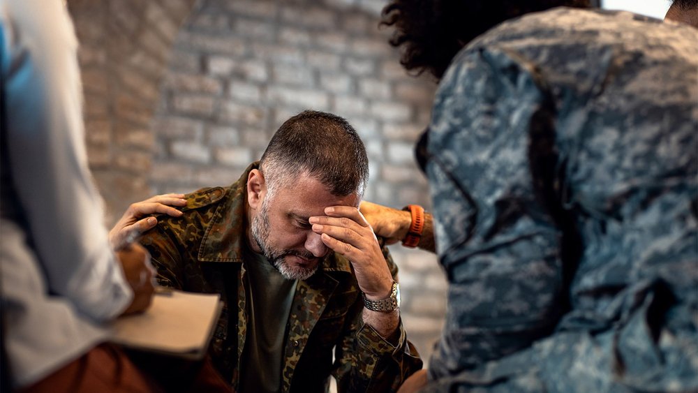 A service member in camouflage is comforted by a hand on the shoulder.
