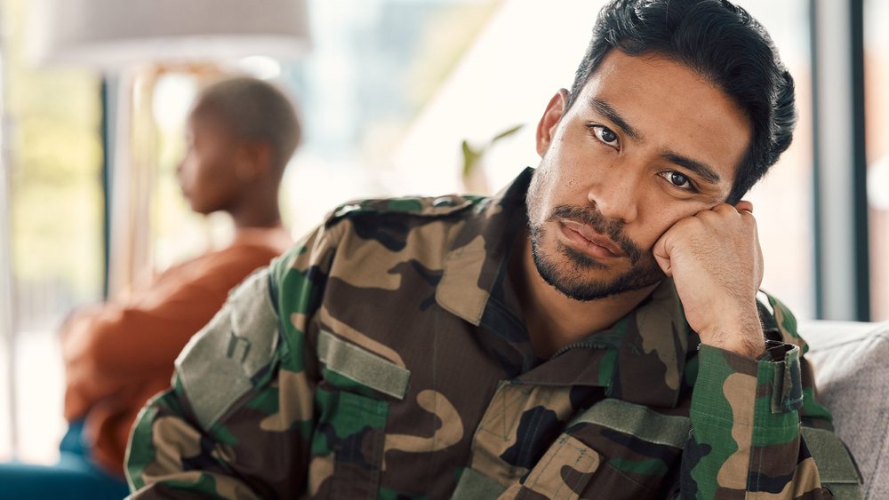 A man in military uniform rests his head on his hand, looking downcast.