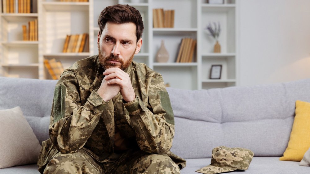 A man in military uniform sits on a couch with hands clasped, looking pensive.
