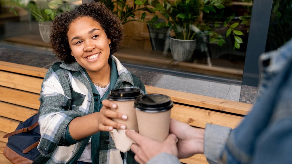 A smiling woman holds two takeout coffee cups while sitting on an outdoor bench.