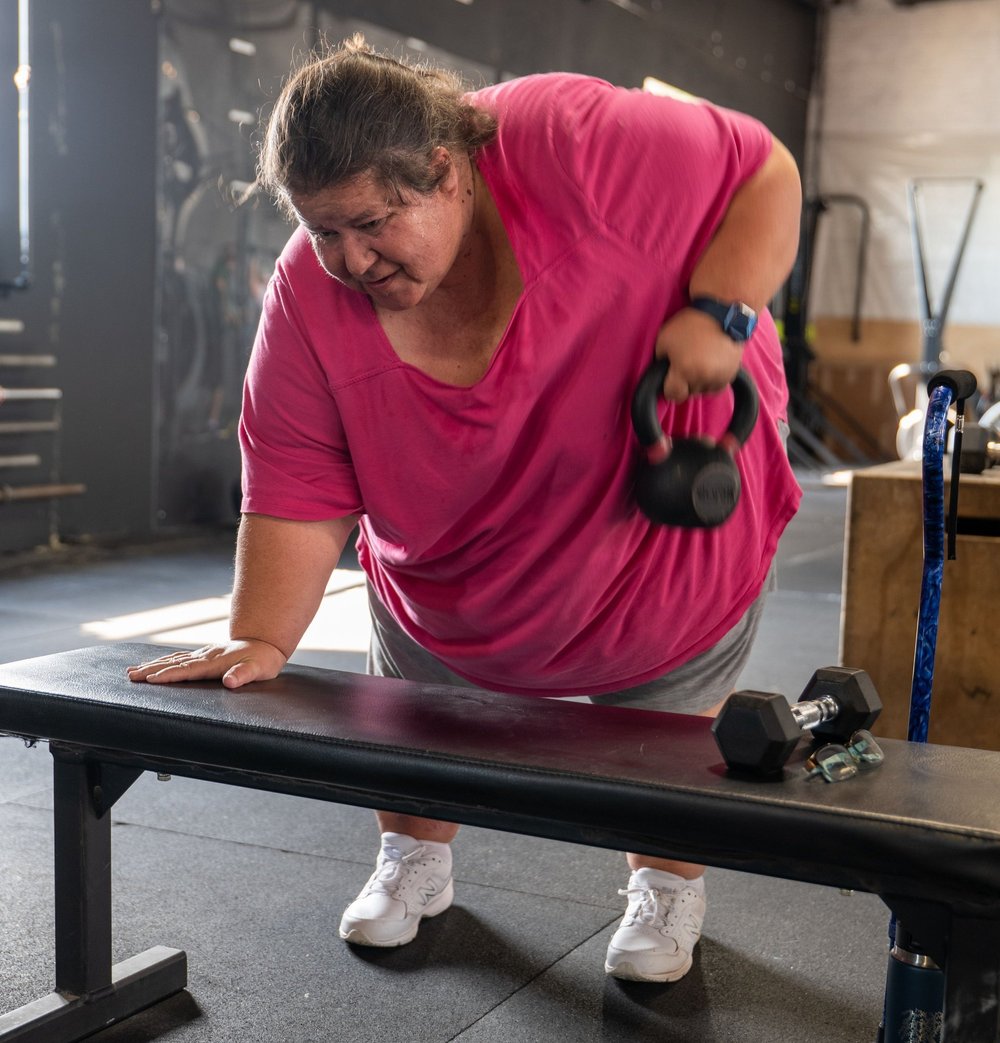 A woman lifts a kettlebell beside a gym bench, with a cane resting nearby.