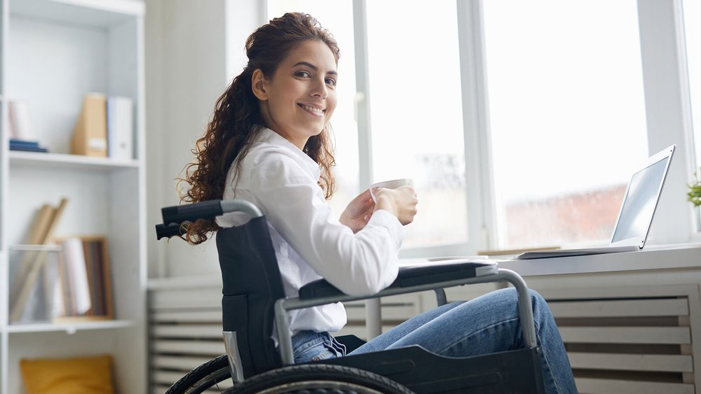 A woman in a wheelchair holds a mug at a desk with a laptop.