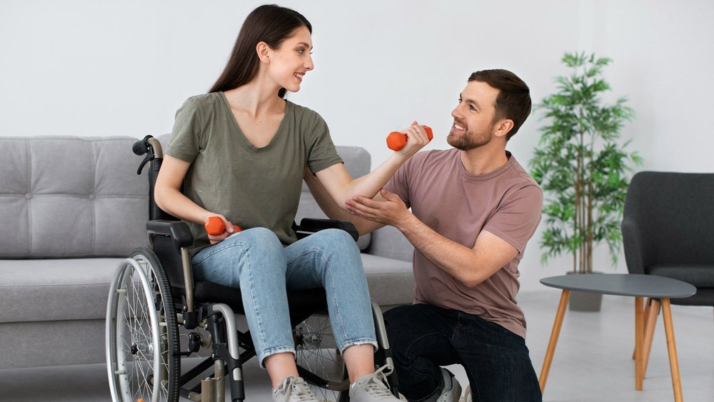 A woman in a wheelchair lifts small dumbbells while a trainer supports her arm.