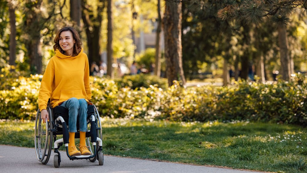 A smiling woman in a wheelchair rolls along a path through a sunlit park.