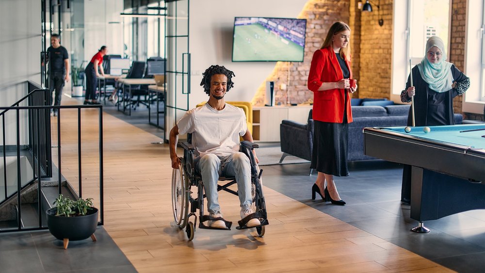 A man in a wheelchair moves through a modern office lounge as others socialize nearby.