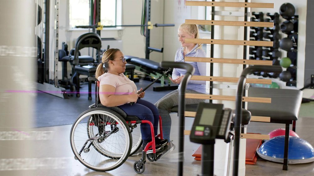 A woman in a wheelchair uses gym equipment while an instructor stands nearby.