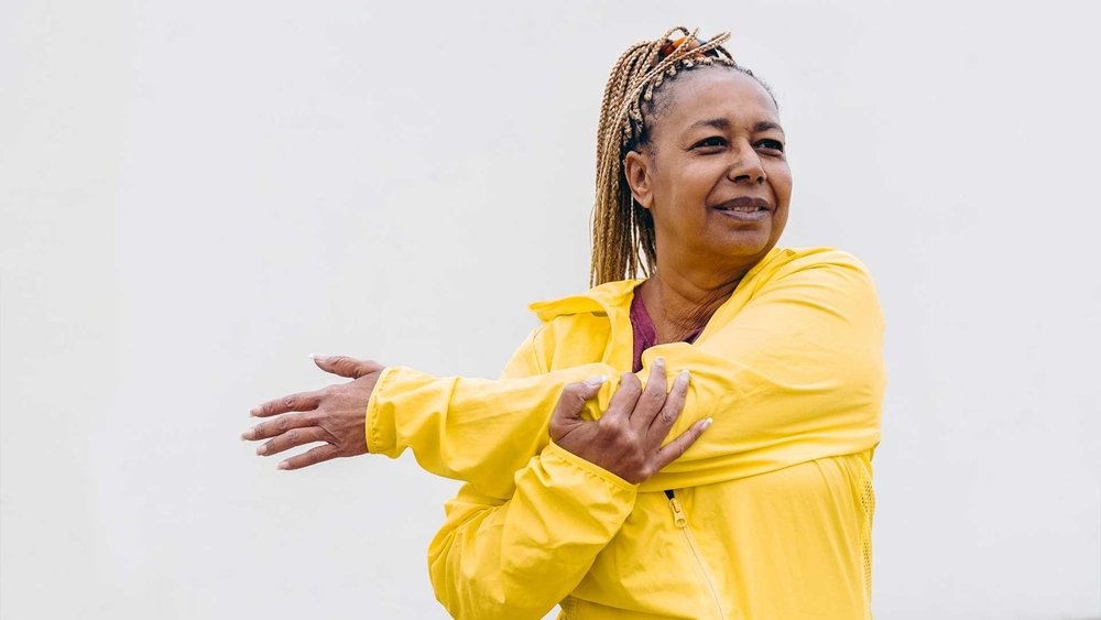 A woman in a yellow jacket stretches one arm across her chest.