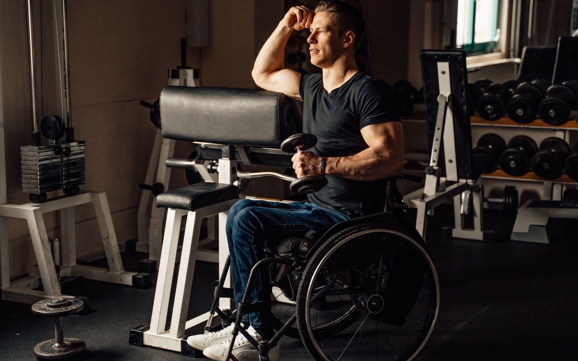 A man in a wheelchair holding a dumbbell beside a gym weight rack.