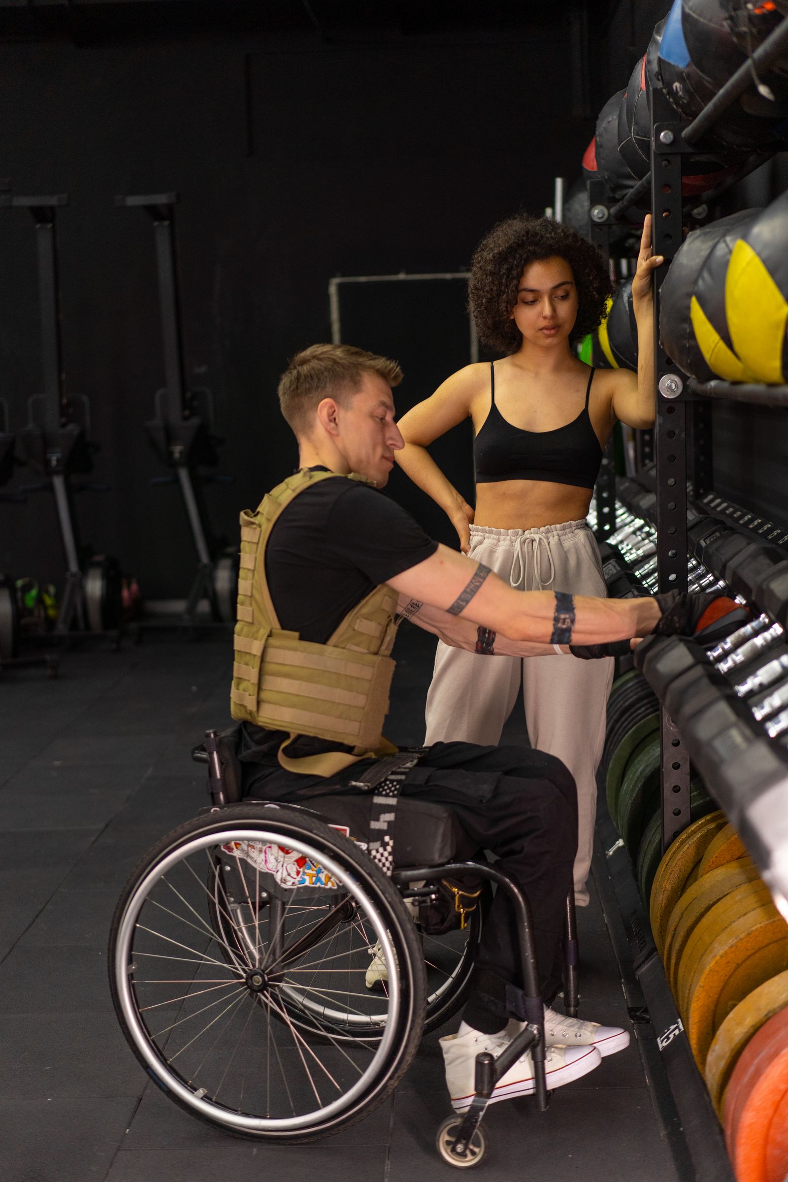 A man in a wheelchair and a training partner beside a rack of medicine balls.